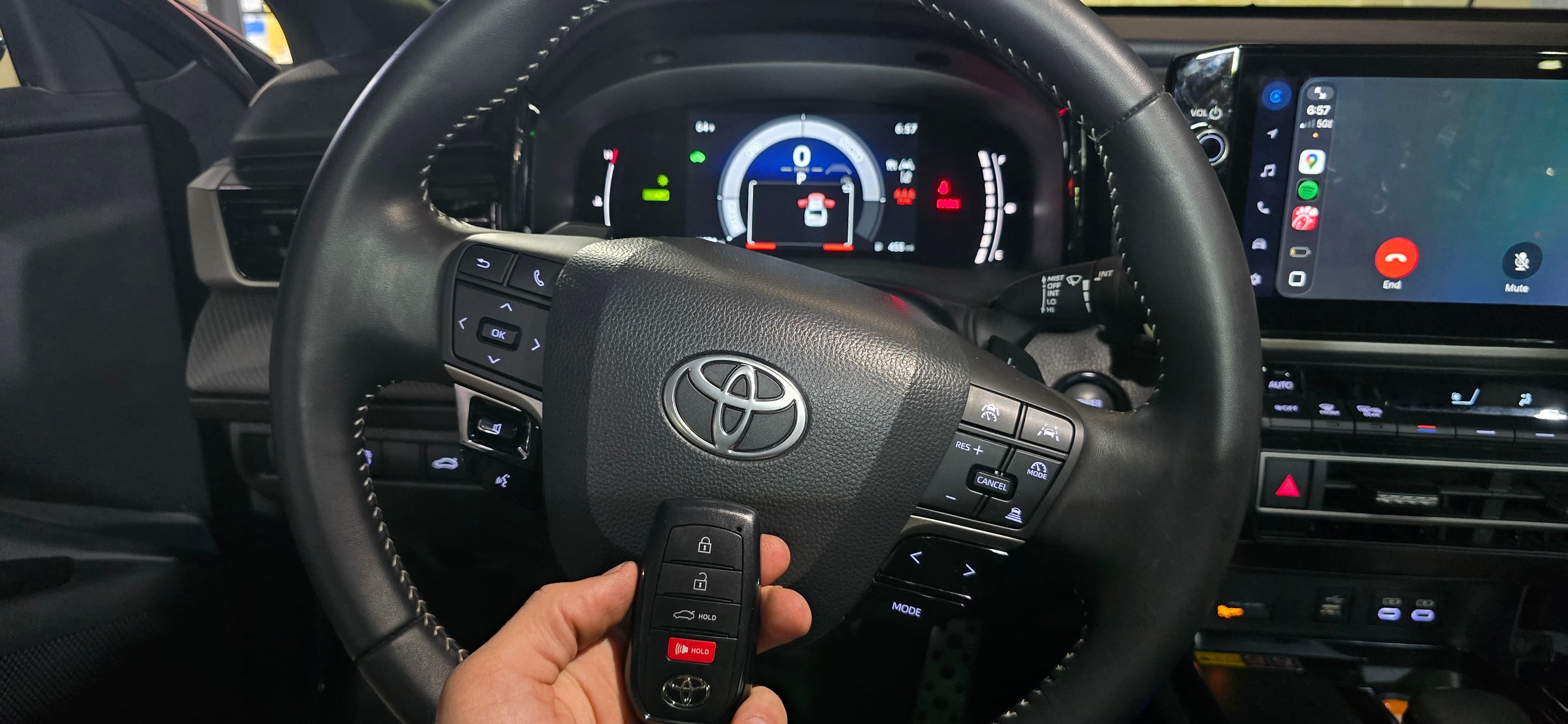 Hand holding a Toyota key fob in front of a steering wheel and digital dashboard.