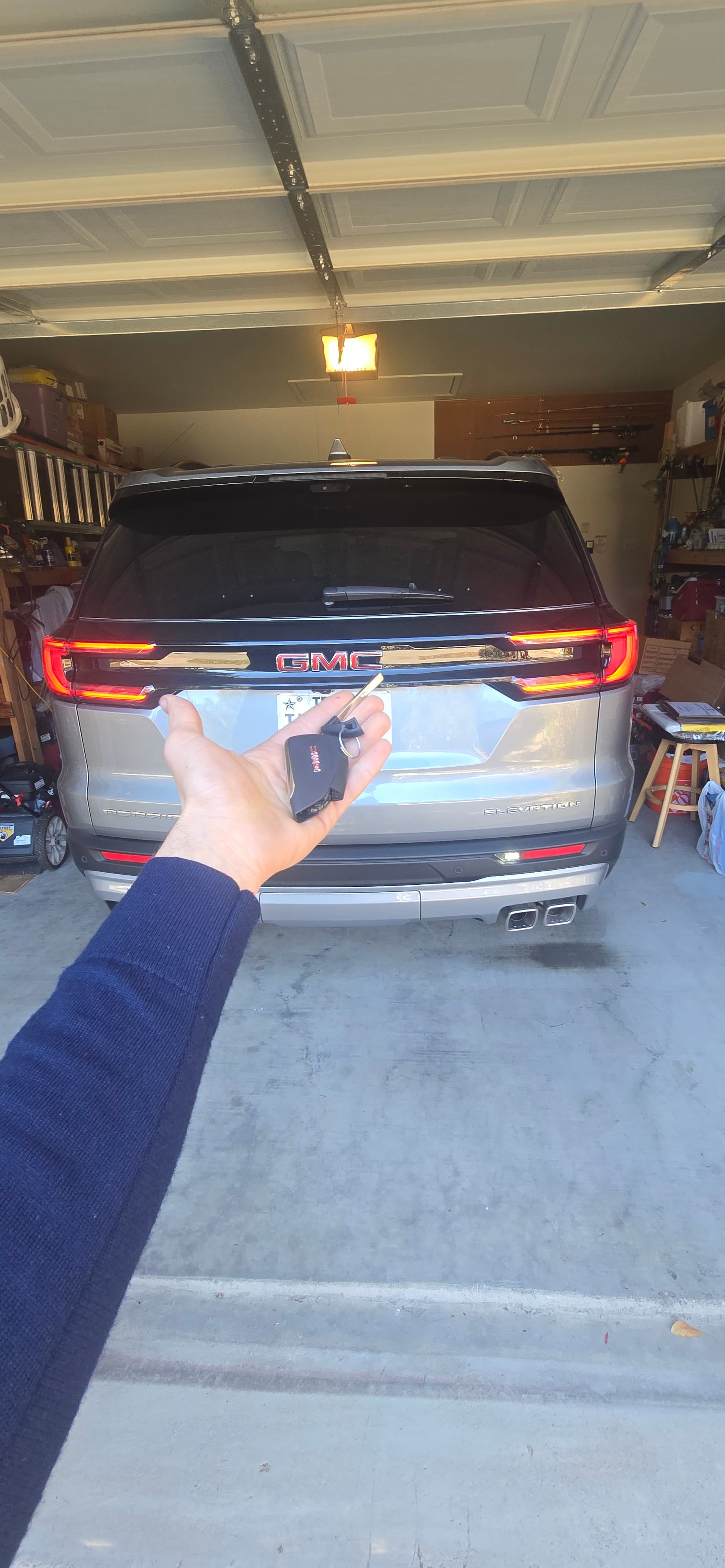Hand holding a GMC key fob in front of a silver SUV with red taillights.