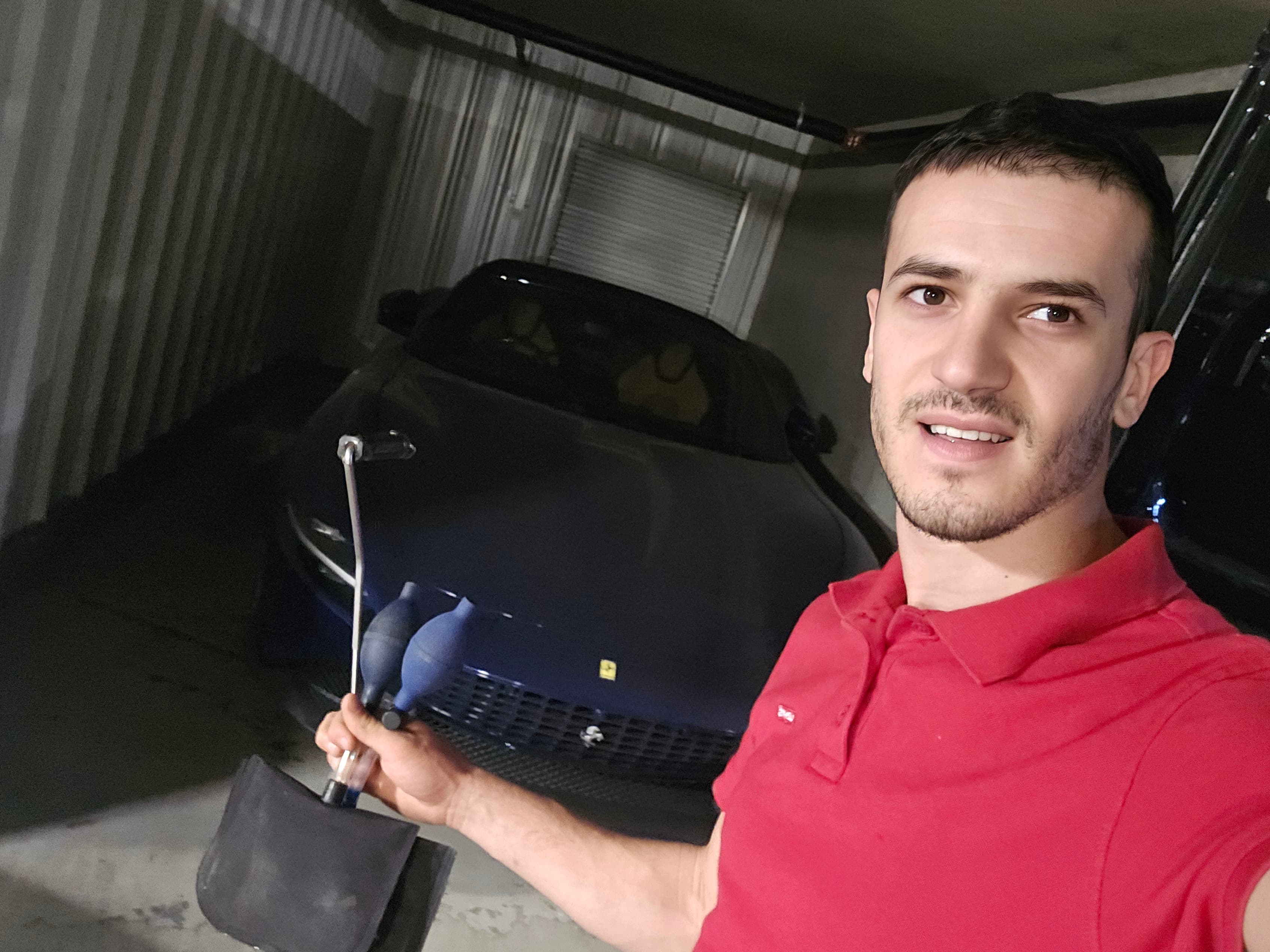 Smiling man in red polo holding an air wedge tool before a blue Ferrari.