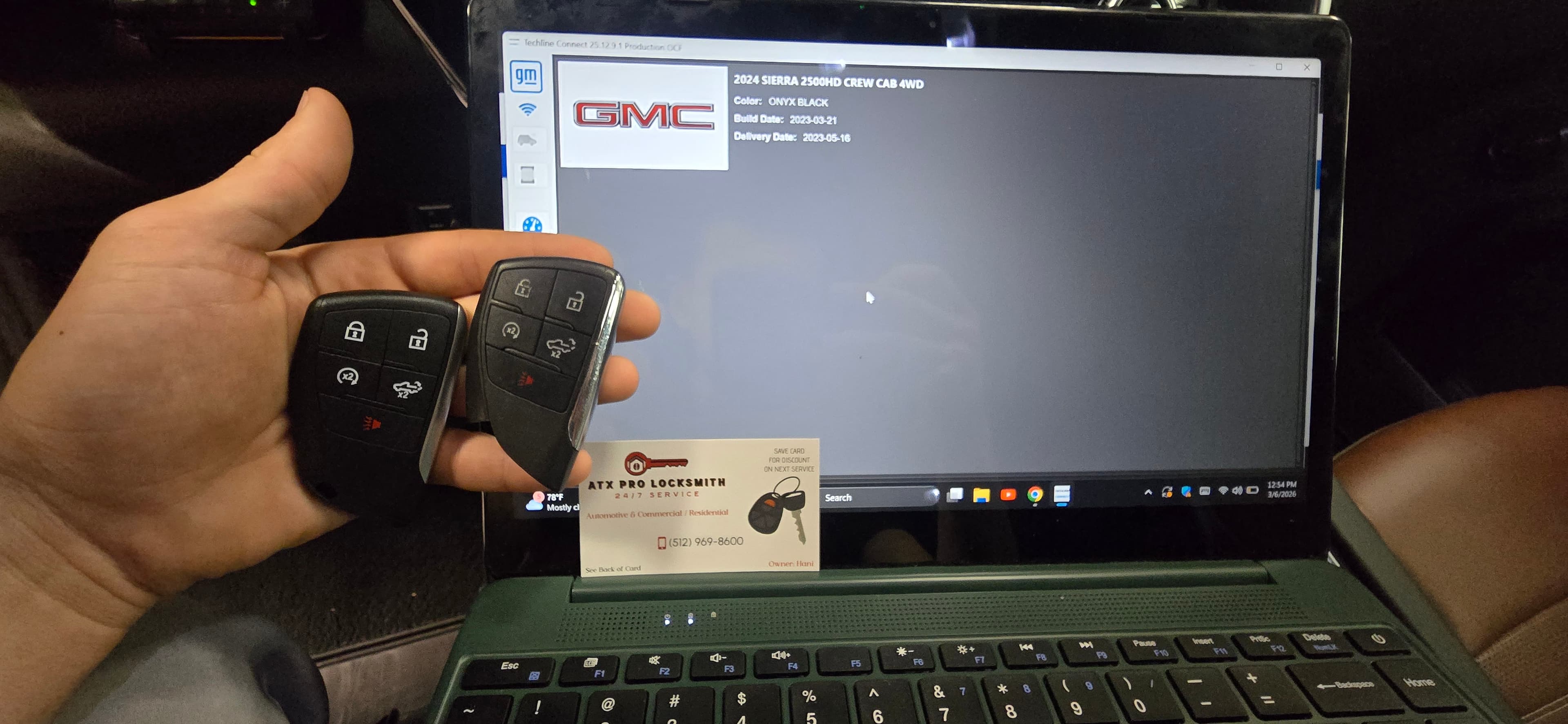 Hand holding two GMC key fobs over a laptop screen and a locksmith business card.